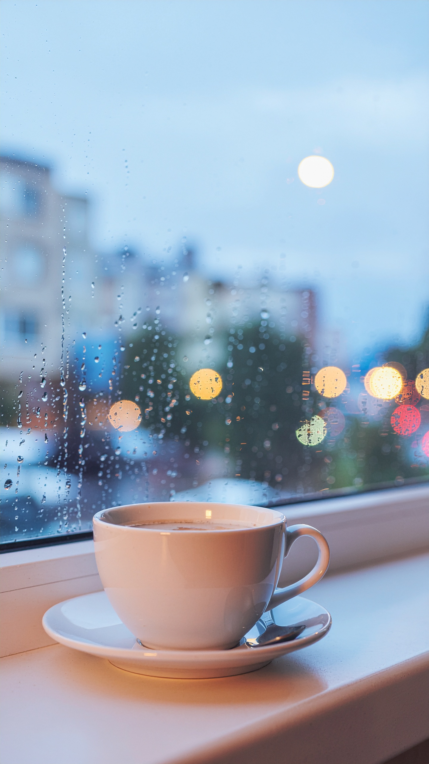 Xícara de café sobre peitoril de janela, com gotas de chuva no vidro e luzes urbanas desfocadas ao fundo, criando uma atmosfera aconchegante e introspectiva. A iluminação suave destaca o branco da porcelana, enquanto o fundo azul acinzentado sugere um dia chuvoso.